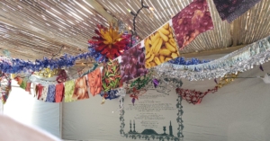 a series of 5” cotton squares for quilting, with fruits and vegetables on them strung together as a garland hanging from a sukkah, with decorative hangings in the background