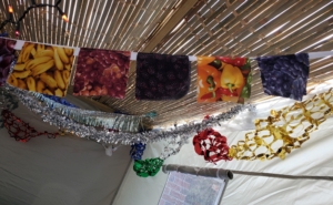 a series of 5” cotton squares for quilting, with fruits and vegetables on them strung together as a garland hanging from a sukkah, with decorative hangings in the background