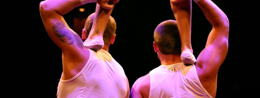 picture of the backs of two gymnasts together supporting a third gymnast on their shoulders (just the legs of the third gymnast are visible)